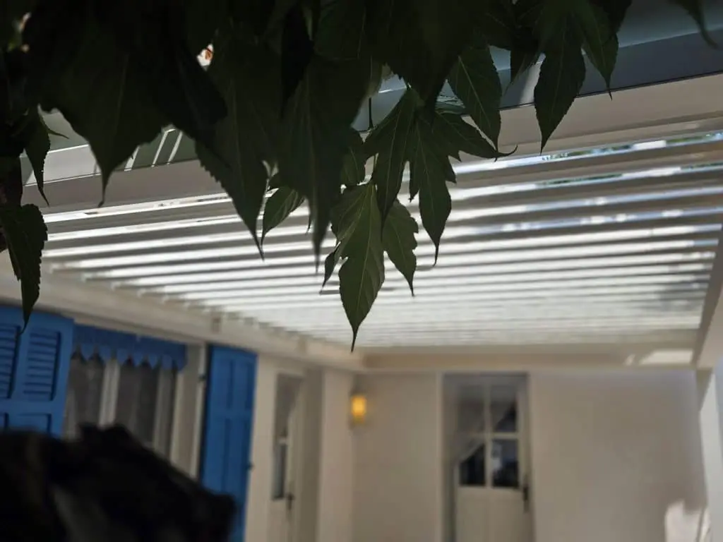 Vue de dessous détaillée d'une pergola bioclimatique adossée à une façade blanche aux volets bleus